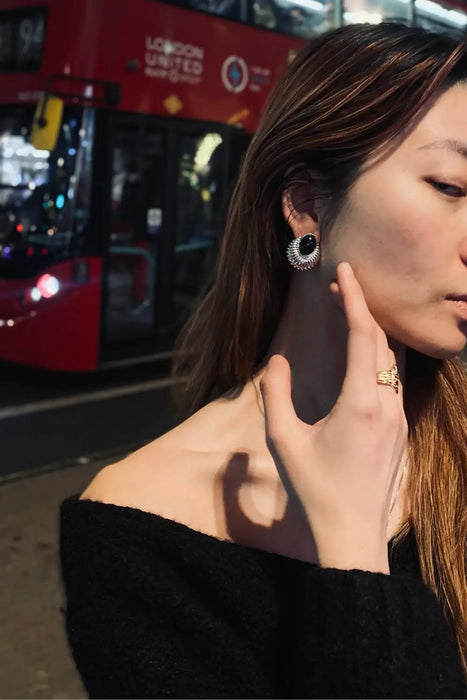 Woman wearing Serenity Black Onyx Silver Earrings from Anaïs & Aimee, standing in front of a classic red London double-decker bus at night. The bold, dark onyx stone contrasts with the surrounding polished silver, capturing a timeless and sophisticated style.