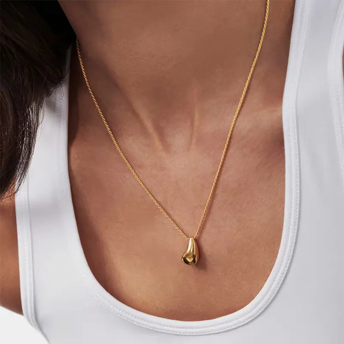The necklace features a slender, delicate chain that sits gracefully on the collarbone. At the center of the chain is a polished, teardrop-shaped gold pendant that subtly catches the light. The background is neutral, emphasizing the gold pendant as it contrasts softly against the woman's skin, showcased against a plain white top. The overall appearance is sophisticated and minimalist, highlighting the necklace as a versatile and stylish accessory.