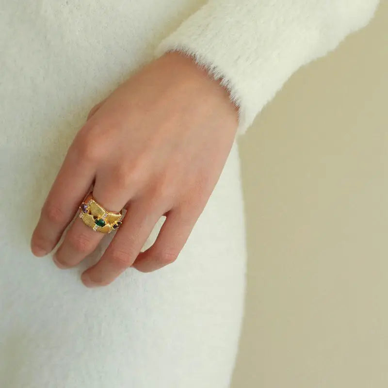 Close-up of a woman's hand wearing the Tempestuous Hues Hoops, featuring an intricate gold-tone design with detailed texturing and vibrant gemstones in green, purple, and red. The woman is dressed in a white outfit, highlighting the elegant and luxurious appeal of these high trending hoop earrings.