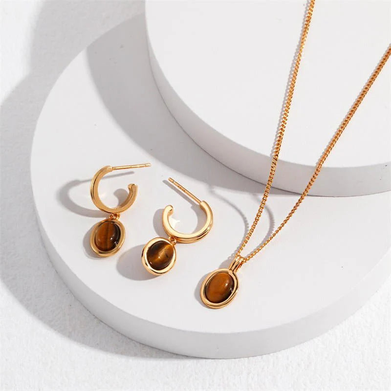 A flat lay photo of a gold-plated jewellery set featuring a pair of hoop earrings and a matching pendant necklace, each adorned with oval-shaped tiger’s eye gemstones. Displayed on white circular platforms, the pieces reflect warm tones and natural stone patterns under soft lighting.