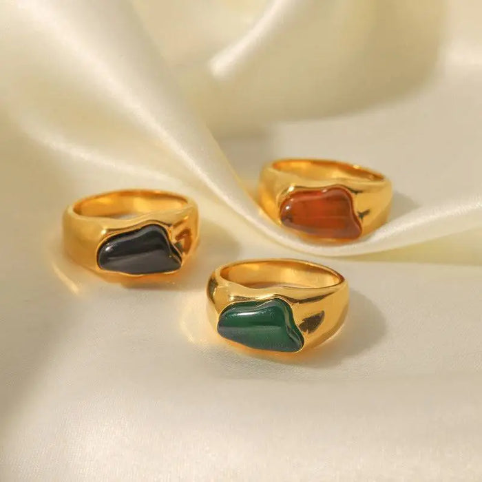 Tri-Color Gemstone Gold Signet Rings displayed on a silky fabric, featuring polished gold-tone bands with vibrant gemstone accents in black, green, and amber hues. These high trending accessories add a touch of bold elegance and sophistication, perfect for making a stylish statement.
