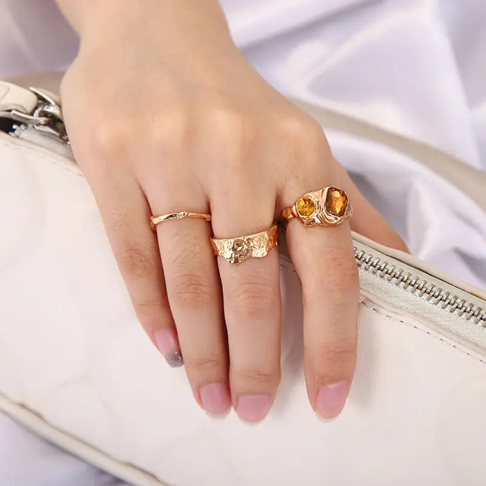 Trio Adjustable Gold Ring Set worn on a hand holding a white clutch - features three unique rings: one with an amber gemstone, a hammered design ring, and a delicate open-ended wavy pattern ring, highlighting their elegant and versatile style.