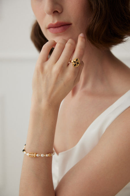 A woman elegantly displaying a vintage-style gold ring with a geometric design on her finger and wearing a matching gold bracelet with pearls on her wrist, set against a soft white background.