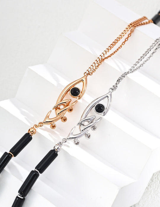 Artistic display of two evil eye necklaces, one with a gold finish and the other silver, each featuring a black gemstone eye and decorative filigree extensions, beautifully arranged on a geometric white stair-step display.