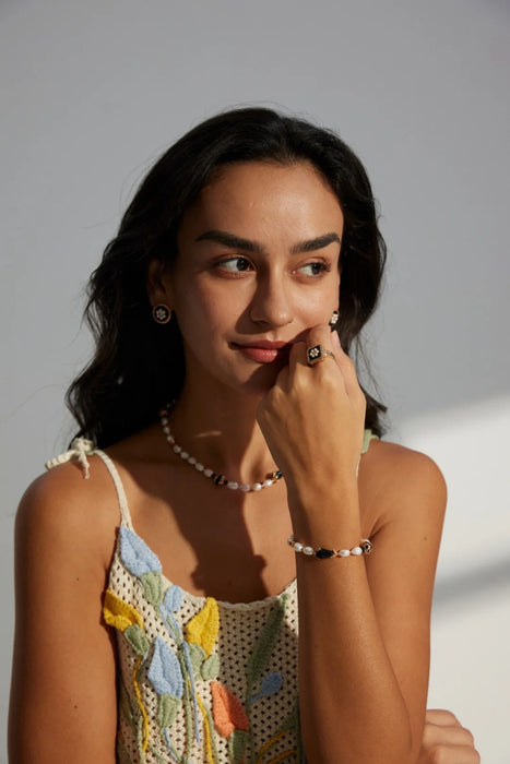 A woman wearing the Vintage Grace Pearl and Enamel Collection, featuring matching black enamel and pearl earrings, a statement ring, a pearl bracelet, and a pearl necklace. The jewellery pieces add a touch of vintage charm and elegance, beautifully complementing her floral crochet top. The model looks thoughtfully to the side, showcasing the coordinated and timeless style.