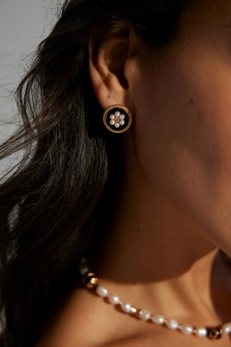 Close-up of a woman wearing the Vintage Grace Pearl and Enamel Earrings. The round gold earrings feature a black enamel background with a delicate floral design made of pearls and a central crystal, exuding vintage elegance. A pearl necklace complements the look, adding a timeless touch to her style.
