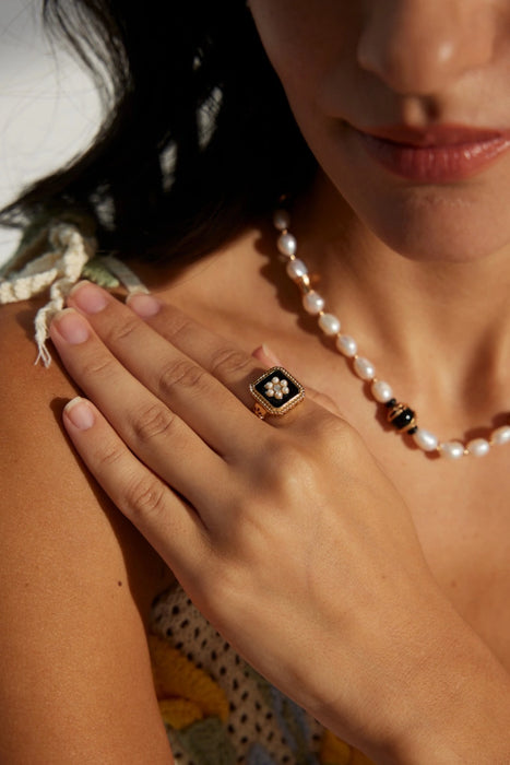 Close-up of a woman wearing the Vintage Grace Pearl and Enamel Ring on her right hand, paired with a pearl necklace. The gold adjustable ring features a black enamel square with a floral pearl and crystal design, exuding vintage charm and elegance against her skin.