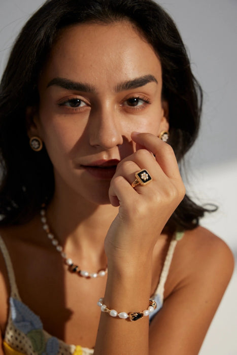 Close-up of a woman wearing the Vintage Grace Pearl and Enamel Ring, along with matching pearl and enamel earrings and a pearl bracelet. The gold ring features a black enamel square with a delicate pearl floral design, adding a touch of elegance to her look. She gazes softly into the camera, showcasing the timeless beauty and sophistication of her jewellery ensemble.