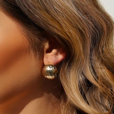 Close-up image of a woman wearing Anais & Aimee Voluminous Gold Hoop Earrings. The earrings feature a textured design and secure butterfly backing, adding a touch of bold elegance to her look.