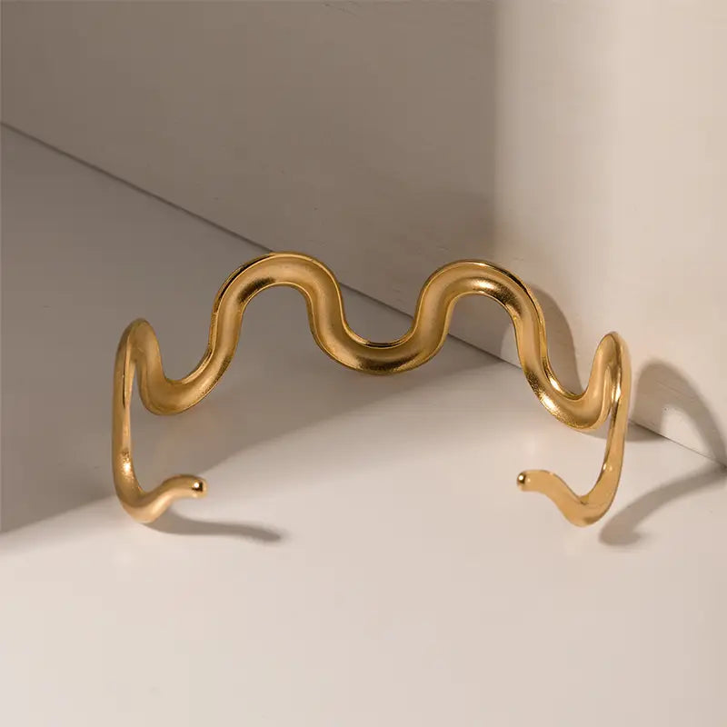 Gold wave-shaped cuff bracelet on white surface with shadow highlighting curved design.