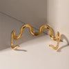 Gold wave-shaped cuff bracelet displayed on a white surface with a shadow cast, showing its curved design.