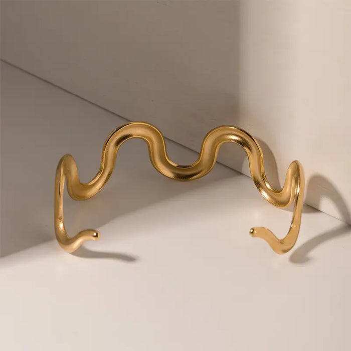 Gold wave-shaped cuff bracelet displayed on a white surface with a shadow cast, showing its curved design.
