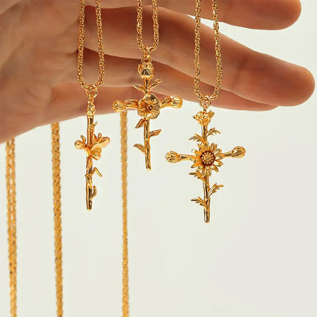  A close-up image showing three gold necklaces, each with a pendant shaped like a cross adorned with different floral designs. The pendants are held by a hand, showcasing their intricate details and craftsmanship. The chains are also gold with a twisted design, complementing the ornate pendants. The background is neutral, highlighting the elegance and beauty of the necklaces.