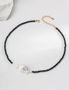 Elegant necklace featuring polished black onyx beads interspersed with subtle golden links, leading to a central large baroque pearl, beautifully showcased on a textured white surface.