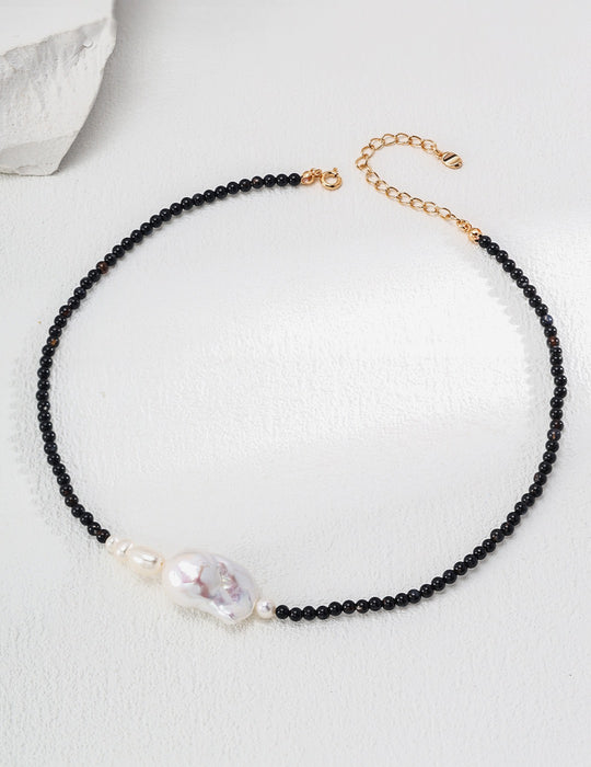 Elegant necklace featuring polished black onyx beads interspersed with subtle golden links, leading to a central large baroque pearl, beautifully showcased on a textured white surface.