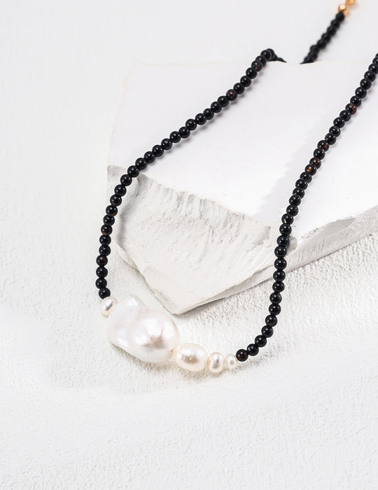 Close-up of a sophisticated black onyx bead necklace with a prominent, irregularly shaped baroque pearl at its center, flanked by smaller pearls, elegantly displayed on a geometric white prop.