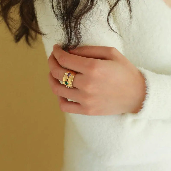 Close-up of a woman's hand wearing the Radiance Gemstone Ring, showcasing an intricate gold-tone design with detailed texturing and vibrant gemstones in green, purple, and red. These high trending hoop earrings add a touch of luxury and elegance, making a stylish fashion statement.