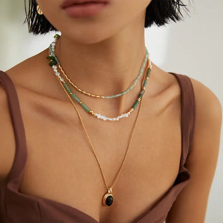 The primary necklace features small green crystals, paired with a golden chain. She also wears a longer gold necklace with a sizable oval black pendant. The overall look is stylish and sophisticated, set against her brown tank top which offers a neutral backdrop, highlighting the jewelry. 
