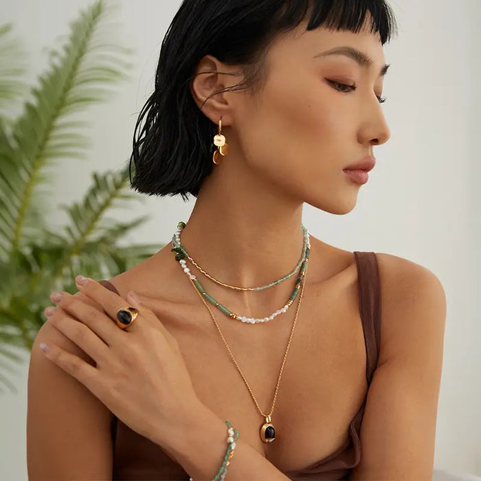 She is wearing a multi-layered necklace combination, one of which appears to be a delicate chain with small green crystals. Additionally, she has a longer gold chain with a black pendant. Complementing her necklaces, she wears matching green crystal bracelets and stylish gold earrings. The setting is serene, possibly indoor, with a hint of tropical plants in the background, emphasizing a fresh and natural aesthetic.